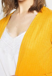 Žlutý žebrovaný kardigan přes bílý top s krajkovým lemem. Kardigan má měkkou texturu a otevřený přední design, který vystavuje vrstvené tkaniny.