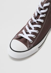 Zapatilla de lona marrón con cordones blancos y punta de goma. Presenta una franja negra a lo largo de la suela y ojales plateados para los cordones.