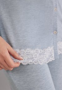 Lichtblauwe stof top met een witte kantafwerking langs de onderrand; beschikt over een knoopsluiting en een gladde textuur. Hand houdt de afwerking vast.