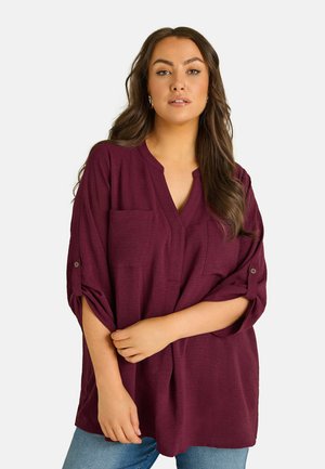 Vrouw met lang bruin haar die een losse bordeauxrode blouse met opgerolde mouwen en blauwe spijkerbroek draagt, staand tegen een effen achtergrond.
