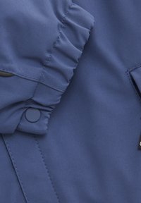 Blå jacka i tyg med strukturerad yta, featuring elastiska rynkade ärmslut och tryckknappsstängning. Inkluderar en tagg på sidan.