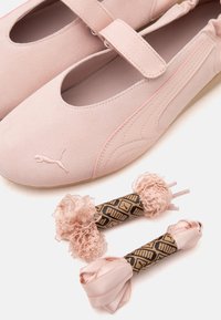 Ballerines en daim rose clair avec une bride, ornées du logo Puma, accompagnées de deux paires de lacets décoratifs roses, dont un volanté, sur fond blanc.
