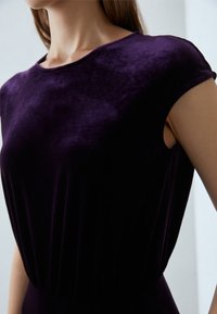 Lila sammetklänning med rundad halsringning och korta ärmar. Tyget har en mjuk textur, och midjan är rynkad för en svängande silhuett.