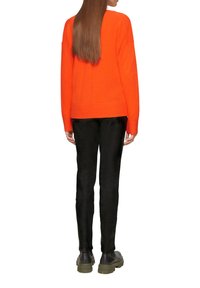 Orangener Pullover mit langen Ärmeln, rundem Ausschnitt und gerippten Bündchen. Kombiniert mit schwarzen Hosen und dunklen Schuhen, von hinten betrachtet.