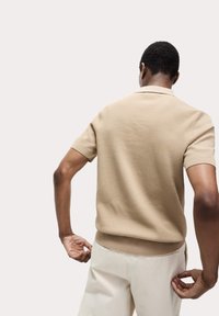 Suéter de polo de punto beige con mangas cortas y puños acanalados, combinado con pantalones color blanco roto. La textura parece suave y un poco ajustada.