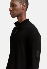 Schwarzer Half-Zip-Pullover mit gerippter Textur, ausgestattet mit einem hohen Kragen und einem Logo-Patch am linken Ärmel. Einfaches und körpernahes Design.