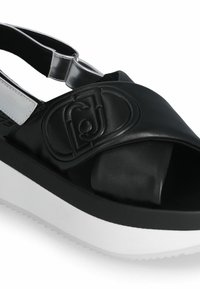 Black leather sandal with wide cross strap, featuring embossed logo, clear elastic heel strap, and thick white platform sole.