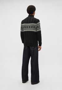 Čierny sveter s vrkočovým vzorom a bielym vzorovaným pásom pri ramenách, doplnený voľnými tmavými džínsami.