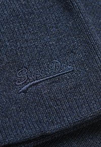 Tejido de punto azul oscuro con una superficie texturizada; un logo "SuperDry" está bordado en un color a juego.