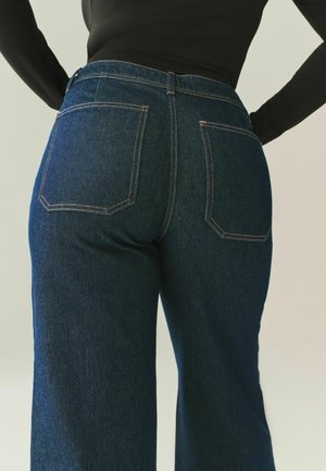 Dark blue denim jeans with a high waist, wide legs, and contrasting orange stitching. Features two back pockets with angular designs.