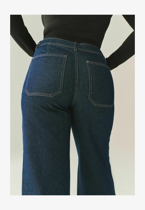 Sötétkék farmernadrág magas derékkal, bő szárú és kontrasztos narancssárga varrással. Két hátsó zseb található rajta, éles szögű mintákkal.