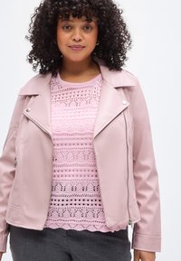 Hellrosa Jacke aus Kunstleder mit notched Revers und Reißverschluss vorne, getragen über einem spitzenbesetzten rosa Oberteil mit filigranen Mustern und einem soliden Ausschnitt.