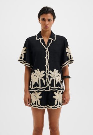 Vrouw draagt zwart T-shirt met korte mouwen en korte broek met crèmekleurige palmboomborduursel en gegolfde randen, staand tegen een witte achtergrond.