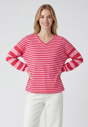 Jonge vrouw lacht met handen in haar zij, draagt een roze- en rood gestreept shirt met lange mouwen en witte broek tegen een effen achtergrond.