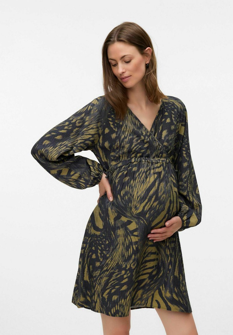 Femme enceinte aux longs cheveux bruns portant une robe longue à manches longues et à motifs, tenant délicatement son ventre d'une main, l'autre main derrière le dos.