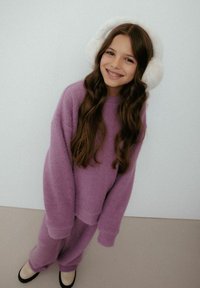 Fioletowy dziergany sweter i pasujące spodnie o fakturowanym wykończeniu. Białe puszyste nauszniki. Tło w jasnoszarym kolorze. Czarne baleriny na stopach.