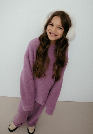 Fioletowy dziergany sweter i pasujące spodnie o fakturowanym wykończeniu. Białe puszyste nauszniki. Tło w jasnoszarym kolorze. Czarne baleriny na stopach.