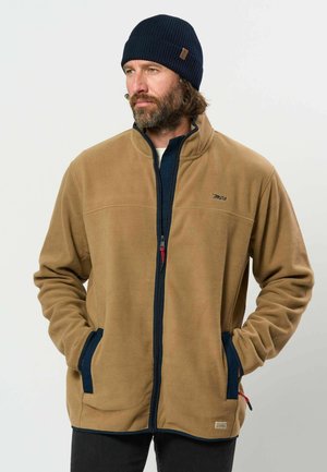 Hombre con barba que lleva una chaqueta polar marrón con cremallera y un gorro de punto negro, con las manos en los bolsillos de la chaqueta, mirando hacia la izquierda.