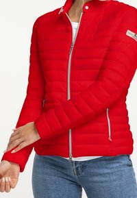 Červená prošívaná bunda s lehkým designem, vybavená stříbrnými zipy a cvočky a dvěma bočními kapsami. Zvýrazněná žebrovanými manžetami.