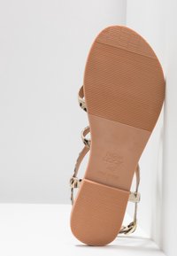 Plochá sandála s hnědým gumovým podrážkem, béžovým a černým vzorovaným svrškem a tenkými pásky. Má strukturovanou podešev pro lepší přilnavost a nastavitelnou přezku.