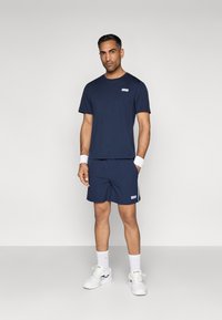 Tamsiai mėlynas sportinis kostiumas, susidedantis iš trumpomis rankovėmis marškinėlių ir šortų, pagamintas iš lygaus audinio, su mažai logotipu ir baltais šonų akcentais.