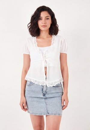 Donna con capelli scuri alla lunghezza delle spalle che indossa una camicetta bianca con bordi in pizzo e una gonna di jeans azzurri chiari, in piedi davanti a uno sfondo semplice.