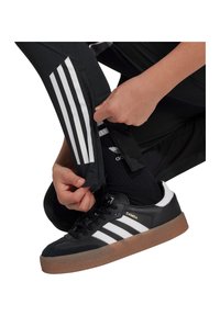 Schwarze Adidas Samba Sneakers mit weißen Streifen und Gummisohle. Die Person passt schwarze Hosen mit weißen seitlichen Streifen und schwarze Socken an.