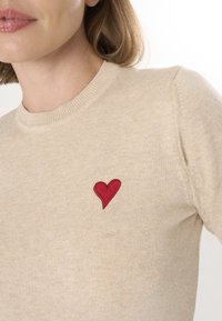 Beige Strickpullover mit einem roten gestickten Herz auf der linken Brust. Die Textur ist weich und hat einen gerippten Halsausschnitt. Einfaches Design.