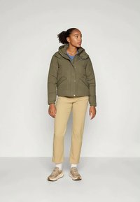 Maslinasto zeleni podstavljeni kaput s kapuljačom, zatvaranjem na zip i bočnim džepovima. Nosite ga preko svijetloplavog košulje i bež hlača, upotpunjeno bež cipelama.