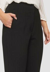 Zwarte, hoge taille broeken van een gladde stof, met zijzakken en scherpe plooien die langs de voorkant lopen voor extra detail.