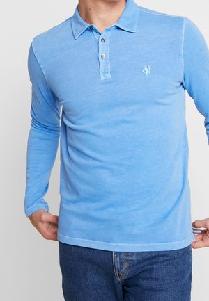 Mand iført en lyseblå poloshirt med lange ærmer, knapper og lille hvidt logo på brystet, kombineret med mørkeblå jeans.