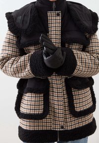 Persona vestida con un abrigo de pata de gallo en marrón y negro con detalles en piel de oveja negra y guantes de cuero negros, con las manos juntas.