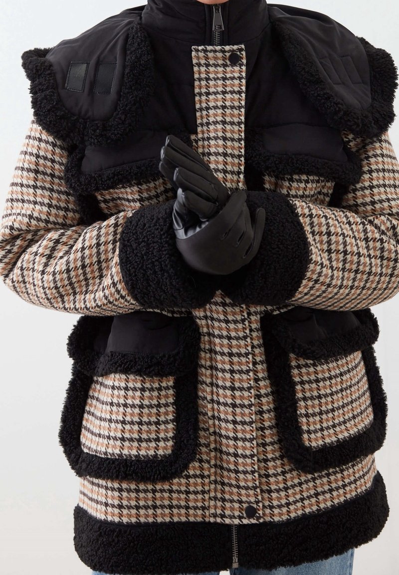 Persona vestida con un abrigo de pata de gallo en marrón y negro con detalles en piel de oveja negra y guantes de cuero negros, con las manos juntas.