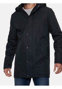 Man i svart huva kappa med knappar framtill över en grå skjorta och blå jeans, som står framför en helt vit bakgrund.