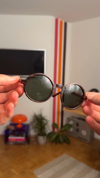 Occhiali da sole rotondi con lenti scure e una montatura marrone maculata sono tenuti in mano, mostrando il design leggero e la texture liscia.