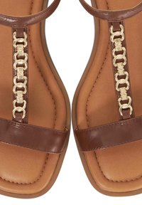 Brown sandals with a flat sole, featuring a gold chain detail at the center and a smooth leather finish. Straps are minimal and elegant.