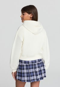 Giovane che indossa una felpa bianca con cappuccio e una gonna a pieghe a quadri blu e neri, di profilo su uno sfondo neutro.