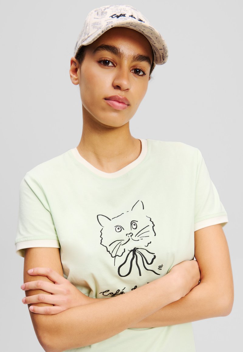 Tricou din bumbac de culoare verde deschis, cu mâneci scurte, prezentând un grafic cu o pisică neagră și o captuseală bej. O pălărie cu un model bej completează ținuta.