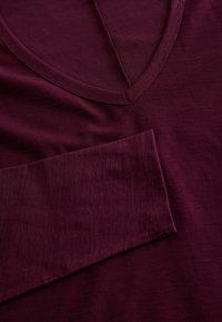 Bordo majica s dugim rukavima koja ima dubok V-izrez, glatku teksturu i ravne šavove. Tkanina izgleda lagano s suptilnim sjajem.