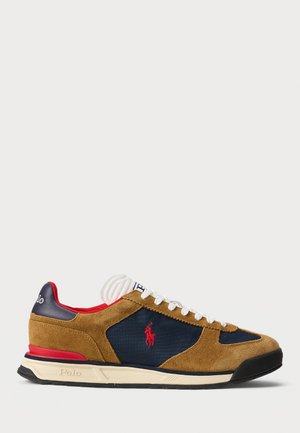 Sneaker in suede marrone con dettagli in tessuto blu navy, lacci bianchi, rifiniture rosse sul tallone e un logo classico sul lato. Suola piatta in gomma nera.