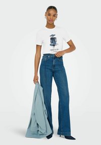 T-shirt bianca con una grafica illustrata e testo, abbinata a jeans blu a vita alta. Il modello tiene un blazer azzurro.