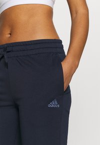 Mano de mujer en el bolsillo de un pantalón deportivo azul marino de Adidas con cintura ajustable, mostrando el vientre y el logo en el muslo sobre un fondo claro.