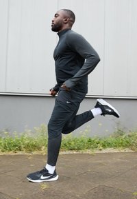 Schwarzer sportlicher Trainingsanzug mit strukturierten Ärmeln, ausgestattet mit einem Reißverschlusskragen und Seitentaschen. Getragen mit schwarzen Laufschuhen und weißen Socken.