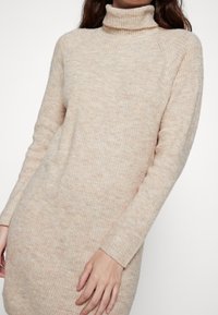 Robe en tricot col roulé en beige doux, featuring un tissu texturé, des manches longues et une silhouette ajustée avec une coupe légèrement décontractée.