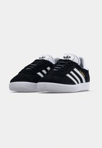 Svarte Adidas Gazelle-sneakers med hvite striper, hvite såler og gullfarget «GAZELLE»-tekst på siden, vist fra frontvinkel.