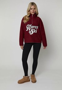 Bordowy polar z półzamkiem i białą grafiką "Apres Ski". Połączony z czarnymi legginsami i beżowymi botkami za kostkę.