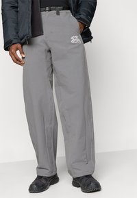 Svetlo sivi zunanji hlače z ohlapnim krojem, z črnim pasom, belim logotipom in vidnim šivom vzdolž šivov.