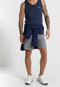 Canotta blu navy con logo, pantaloncini sportivi grigi e sneakers bianche. Una felpa blu navy è legata in vita. La pelle è abbronzata e le braccia sono tatuate.