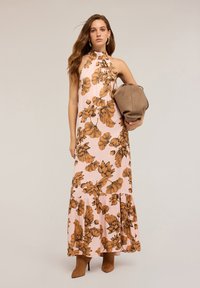 Rochie maxi dintr-un material roz deschis, �împodobită cu modele florale maronii, cu decolteu halter și tiv cu volane. Modelul ține o geantă bej.