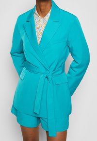 Blazer turchese con cintura a cravatta, caratterizzato da un colletto a scialle e due tasche frontali, indossato sopra una camicia a bottoni floreale azzurra chiara.